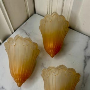 May include: Three vintage, amber-colored glass light shades. Each shade has a scalloped top with a swirl design and a tapered, ribbed body. The shades are arranged on a marble-patterned surface.