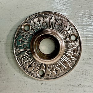 May include: A silver-toned metal door knob rosette with a decorative design featuring a repeating pattern of stylized flowers and leaves.
