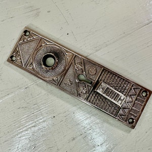 May include: A silver-toned door plate with an intricate geometric design. The plate has a keyhole and a round knob.