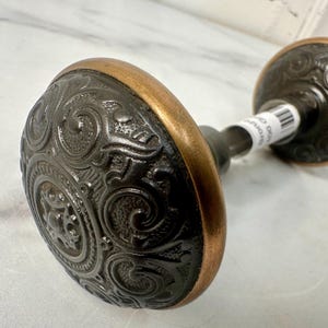 May include: An antique bronze door knob with an ornate, swirling pattern. The knob has a dark finish with a copper-colored rim. The door knob is a vintage style and is likely used for interior doors.