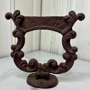 May include: Ornate, antique-style cast iron wall hook with a dark reddish-brown finish. The hook features a decorative, scroll-like design with a rectangular space in the center. The base is round with a single screw hole.
