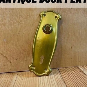 May include: Antique gold door plate with a keyhole and a round knob. The plate has a decorative design around the edges.