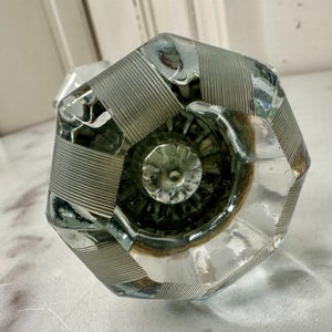 May include: Clear glass octagonal door knob with etched vertical lines and a floral design in the center. The knob has a reflective surface and is likely vintage or antique.