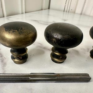 May include: Antique brass and black cabinet knobs with a threaded metal tool. The knobs have an oval shape and a decorative base. The tool is rectangular with a ridged surface. Hardware for furniture restoration.