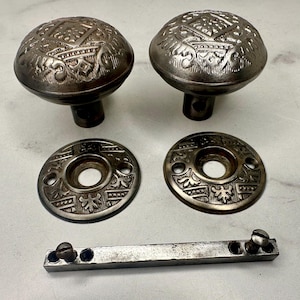 May include: Two antique silver-toned door knobs with ornate, raised patterns. The set includes two matching round backplates and a rectangular metal piece with screw holes. The hardware is likely vintage and shows signs of wear.