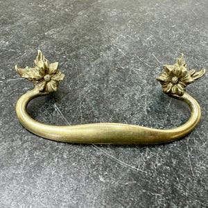 May include: A brass drawer pull with a curved handle and floral accents. The decorative hardware features a curved handle with a flower design on each end. The brass finish gives it a vintage look.