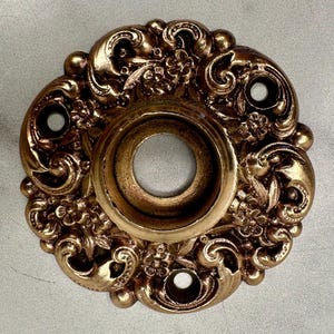 May include: Ornate, antique-style bronze door hardware. The circular piece features a central hole, surrounded by floral and scrollwork details. Three smaller holes are evenly spaced around the perimeter. The finish has a warm, aged appearance.