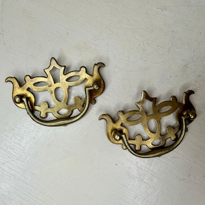 May include: Two antique brass drawer pulls with ornate, openwork designs. Each pull features a curved handle and decorative flourishes, with a warm, aged patina. The pulls are likely for a cabinet or dresser.