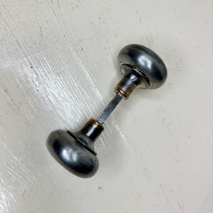 May include: Antique metal door knob with a dark, aged finish. The knob has an oval shape on each end, connected by a rectangular rod. The metal has a slightly tarnished appearance, suggesting age and use. A classic hardware piece.