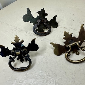 May include: Three antique bronze drawer pulls with a leaf design. Each pull features a decorative backplate and a curved handle. The pulls are arranged on a white surface, showcasing their intricate details and aged patina.
