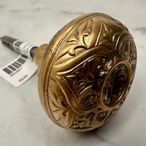 May include: Ornate antique brass door knob with detailed floral and leaf patterns. The knob has a rounded shape and a warm, golden hue. A metal stem extends from the knob, with a tag attached. The knob is likely for a vintage or classic door.
