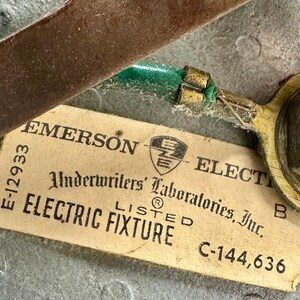 May include: Close-up of an Emerson Electric Fixture tag. The tag is beige with black text, including "EMERSON ELECTRIC" and "Underwriters' Laboratories, Inc." A green wire is attached to a metal component. The background is a metallic surface.