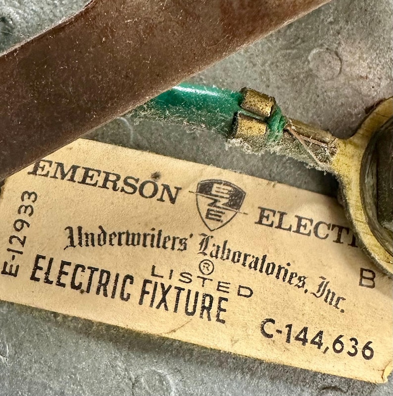 May include: Close-up of an Emerson Electric Fixture tag. The tag is beige with black text, including "EMERSON ELECTRIC" and "Underwriters' Laboratories, Inc." A green wire is attached to a metal component. The background is a metallic surface.