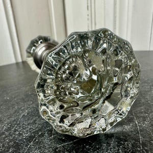 May include: A clear, faceted glass doorknob with a silver-toned metal base. The knob has a textured, irregular surface, reflecting light. The design is vintage, suitable for interior decor.