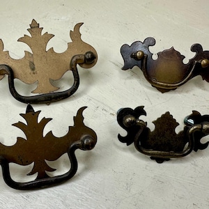 May include: Four antique brass drawer pulls with ornate, floral-inspired designs. Each pull features a dark, curved handle and decorative backplate. The pulls are in varying shades of brown and gold, with a distressed finish.