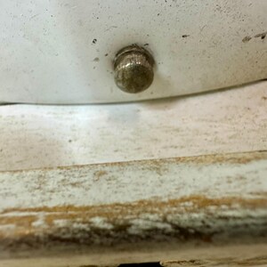 May include: Close-up of a small, tarnished metal knob on a white surface. The knob is round with a textured surface. Below the white surface is a section of distressed, light-colored wood.