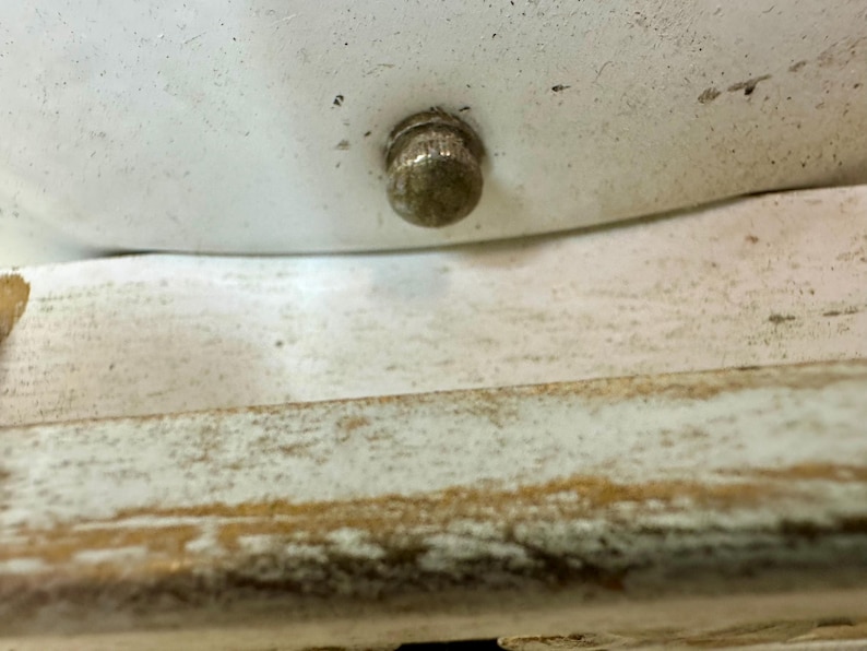 May include: Close-up of a small, tarnished metal knob on a white surface. The knob is round with a textured surface. Below the white surface is a section of distressed, light-colored wood.
