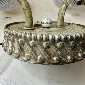 May include: An antique wall sconce with a distressed, off-white finish. The sconce features a decorative base with an ornate, scroll-like design. The curved arms support glass shades. Electrical wiring is visible.