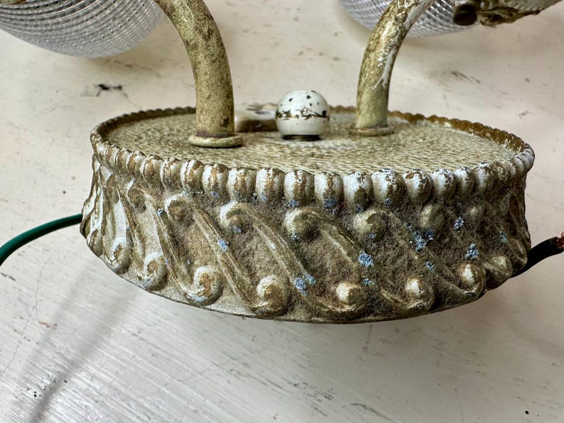 May include: An antique wall sconce with a distressed, off-white finish. The sconce features a decorative base with an ornate, scroll-like design. The curved arms support glass shades. Electrical wiring is visible.