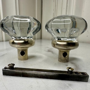 May include: Two clear glass door knobs with a ribbed design, mounted on brass-colored bases. A dark metal plate with screws is also present, likely for installation. The knobs have a vintage aesthetic.