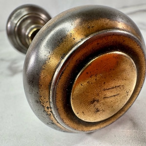 Puede incluir: Un pomo de puerta vintage con acabado en latón y base plateada. El pomo tiene una forma redondeada y una superficie lisa.