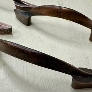 May include: Close-up of several copper-colored cabinet pulls. The pulls have a curved, ergonomic design with a textured finish. The hardware is likely for kitchen or bathroom cabinets, adding a rustic or industrial aesthetic.