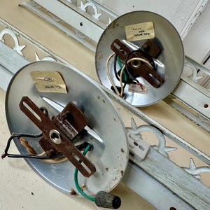 May include: Two vintage light fixtures with metal mounting plates and wiring. The fixtures have a distressed, aged appearance. Decorative white metal trim pieces with a repeating pattern are also present. The fixtures are likely for wall mounting.
