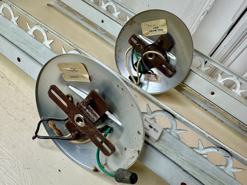 May include: Two vintage light fixtures with metal mounting plates and wiring. The fixtures have a distressed, aged appearance. Decorative white metal trim pieces with a repeating pattern are also present. The fixtures are likely for wall mounting.