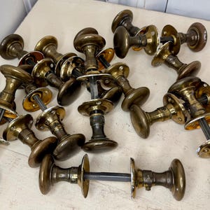 May include: A collection of antique brass door knobs with a tarnished finish. The knobs have a classic mushroom shape and are arranged on a light-colored surface. The hardware includes the knob, a decorative base, and a connecting rod.