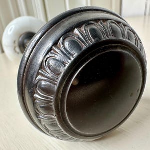May include: Close-up of a vintage door knob with a dark, ornate metal design. The knob features a detailed, textured pattern around the edge and a smooth, dark center. A white porcelain knob is visible in the background.