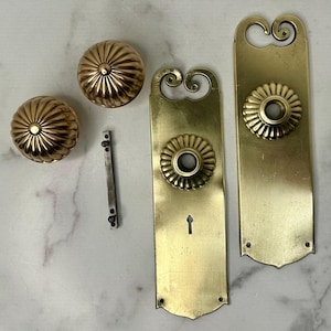 May include: Antique brass door hardware set. The set includes two ornate door knobs, two long backplates with decorative floral accents, and a latch. The hardware has a warm, golden hue and shows signs of age.