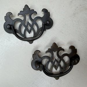 May include: Two antique-style drawer pulls with a dark, aged finish. Each pull features an ornate, openwork design with a curved handle. The pulls are made of metal and are likely for furniture restoration or decorative use.