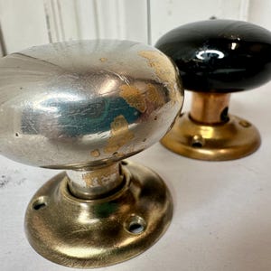 May include: Two antique door knobs. One is silver-toned with wear and tear, the other is black with a brass base. The knobs have an oval shape and are mounted on a round base with screw holes. The door knobs are vintage and show signs of age.
