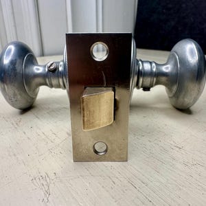 May include: A vintage door knob set with a silver finish. The set includes two rounded knobs and a rectangular plate with a brass-colored latch. The hardware is designed for a door and is likely made of metal.