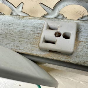 May include: Close-up of a vintage white electrical outlet with a metal screw, mounted on a distressed, light blue wooden surface. Decorative metal accents in a curved design are visible above the outlet.