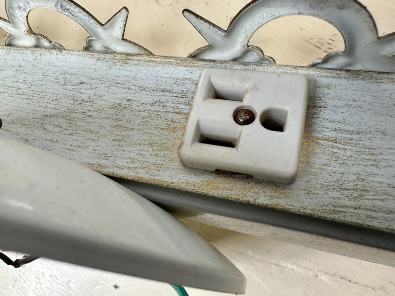 May include: Close-up of a vintage white electrical outlet with a metal screw, mounted on a distressed, light blue wooden surface. Decorative metal accents in a curved design are visible above the outlet.