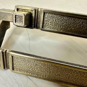 May include: Close-up of a bronze-colored door pull with a textured surface. The rectangular handle has a square accent piece and a decorative border. The hardware is likely for a cabinet or door.