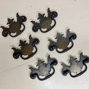 May include: Six antique bronze drawer pulls with a leaf design. Each pull has a decorative backplate and a curved handle. The pulls are arranged on a white surface, showcasing their aged patina and intricate details.