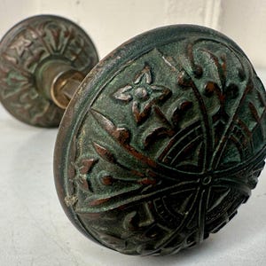 May include: Close-up of two antique bronze door knobs. The knobs have an aged, verdigris finish with intricate floral and geometric patterns. The design includes stylized flowers and leaf motifs, showcasing a vintage aesthetic.