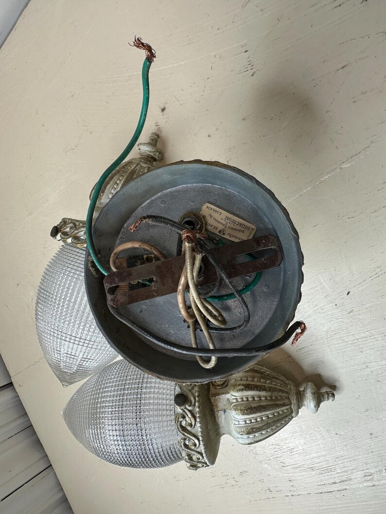 May include: An antique ceiling light fixture with two glass shades. The fixture has a metal base and decorative, off-white accents. Exposed wiring and a green ground wire are visible.