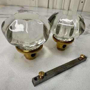 May include: Two clear glass cabinet knobs with a ribbed design and brass bases. A metal bar with two brass screws is also present. The knobs have a vintage aesthetic.