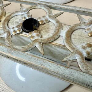 May include: Close-up of a decorative light fixture with a distressed white and gold finish. The fixture features ornate floral and geometric designs, with a black central element. The fixture is mounted on a white surface.