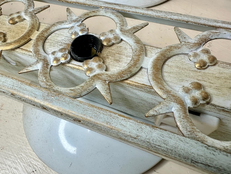 May include: Close-up of a decorative light fixture with a distressed white and gold finish. The fixture features ornate floral and geometric designs, with a black central element. The fixture is mounted on a white surface.