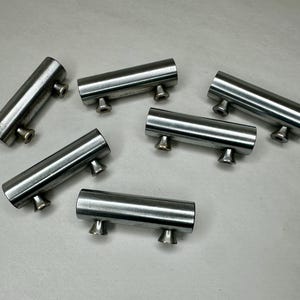 May include: Six brushed nickel cabinet pulls. Each pull is a cylindrical bar with two small, cone-shaped feet. The hardware is arranged on a light gray surface, showcasing their sleek, modern design. These pulls are ideal for updating kitchen or bathroom cabinets.