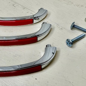 May include: Three chrome-finished cabinet handles with a red accent, along with several silver screws. The handles have a curved design and are likely for furniture or cabinetry. The screws are of varying lengths.