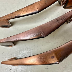 May include: Close-up of several vintage copper-toned cabinet pulls. The pulls have a sleek, angular design with a slightly curved handle. The copper finish shows signs of age and wear, adding to their antique charm. These pulls are ideal for adding a touch of retro style to furniture.