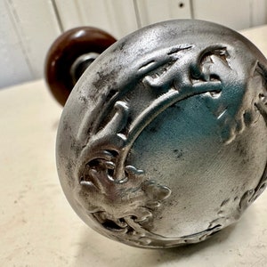 May include: A silver door knob with an ornate design. The knob is round and has a raised design of leaves and flowers.