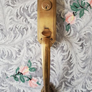 May include: A gold-toned door handle with a decorative design and a keyhole. The handle is attached to a door with a floral pattern.