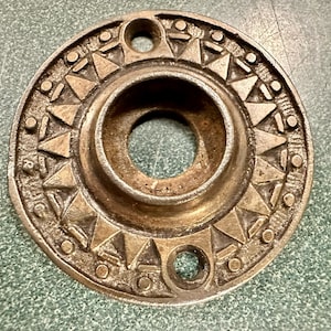 May include: A circular, brass door knob escutcheon with a decorative, geometric design. The escutcheon has a central hole for the knob.