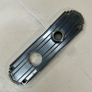 May include: A silver door plate with a rectangular shape and a decorative design of vertical lines. The plate has a round hole in the center and a smaller round hole near the top.